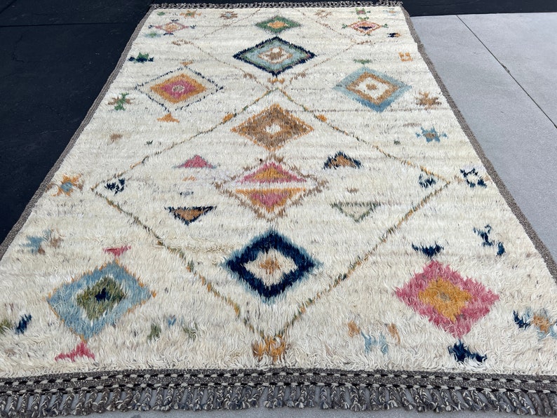 May include: A rectangular, off-white rug with a geometric diamond pattern. The design incorporates various colors, including blue, pink, yellow, and brown. The rug has a textured, shaggy appearance and a fringe border. The rug's dimensions are not visible.