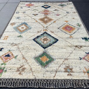 May include: Cream-colored rectangular rug with a geometric diamond pattern in various colors. The design includes diamonds, triangles, and floral motifs in shades of blue, pink, green, and brown. The rug has a fringe border.