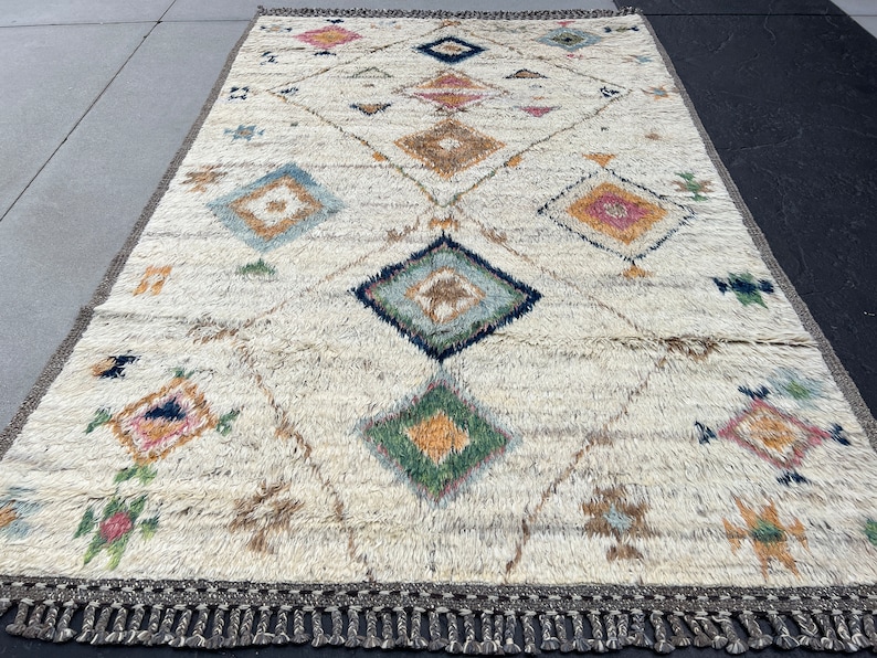 May include: Cream-colored rectangular rug with a geometric diamond pattern in various colors. The design includes diamonds, triangles, and floral motifs in shades of blue, pink, green, and brown. The rug has a fringe border.