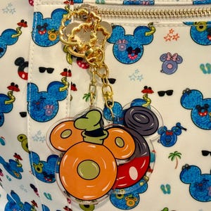 May include: A gold-coloured keychain with a clear acrylic charm featuring a cartoon character design. The charm has orange, green, red, and black details. The background is a white fabric with a blue cartoon character pattern and a gold zip.