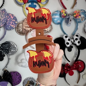 May include: A brown headband holder with two honey pot designs, each with the word "Hunny" in black. The holder is displayed against a wall adorned with various decorative headbands.