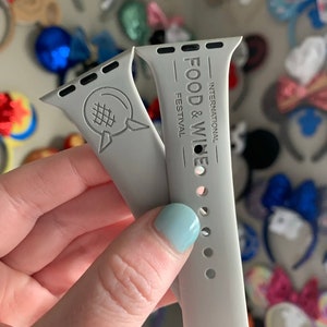May include: Two grey Apple Watch bands with different designs. One band features the Epcot International Food & Wine Festival logo, and the other band has a stylised Epcot logo.