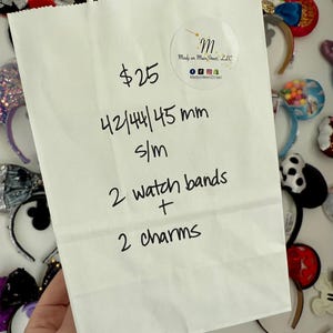 May include: A white paper bag with handwritten text and a circular sticker. The text includes "$25", "42/44/45 mm", "s/m", "2 watch bands + 2 charms". The sticker reads "Mady on Main Street, LLC". The bag is surrounded by decorative headbands.