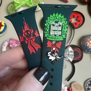 Puede incluir: Dos correas de Apple Watch verdes con diseños diferentes. Una correa presenta un diseño de Haunted Mansion rojo y negro con una calavera y un castillo. La otra correa presenta un diseño de Haunted Mansion verde y blanco con una corona y el texto "The Haunted Mansion Holiday Nights".
