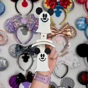 May include: A white Mickey Mouse ghost-shaped headband holder with black ears and a smiling face. The holder is held in front of a wall of various Mickey Mouse ear headbands in different colors and designs.