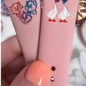 May include: Two pink silicone watch bands with different designs. One band features a cartoon image of three cats, while the other band features a cartoon image of two geese.
