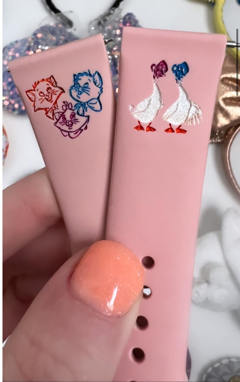 May include: Two pink silicone watch bands with different designs. One band features a cartoon image of three cats, while the other band features a cartoon image of two geese.
