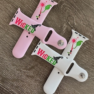 Puede incluir: Dos correas para Apple Watch con un diseño verde y negro de la Bruja Mala del Oeste. Una correa es rosa y la otra es blanca.