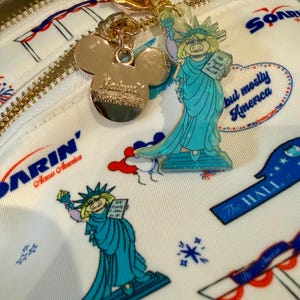 May include: White backpack with a gold zipper and a repeating pattern of red, white, and blue patriotic imagery. The backpack features a gold Mickey Mouse charm and a Statue of Liberty character charm. The text "but mostly America" is visible.