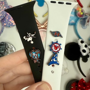 May include: Black and white Apple Watch bands with decorative pins. The black band has a Zero pin and a Jack and Sally heart pin. The white band has a Disney Cruise Line pin, a Mickey Mouse wheel pin, and a Sorcerer Mickey hat pin.