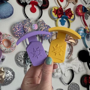 May include: Two purple and yellow sun-shaped ear headbands holders. The holders are made of a soft, flexible material and have a raised sun design. The holders are being held in a hand.