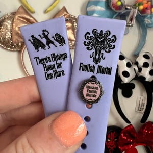 May include: Two purple silicone watch bands with black text and images. The band on the left says "There's Always Room for One More" and has a silhouette of three ghosts. The band on the right says "Foolish Mortal" and has a silhouette of a ghost with a swirling design. A small silver charm with pink text that says "Welcome Foolish Mortals" is attached to the right band.