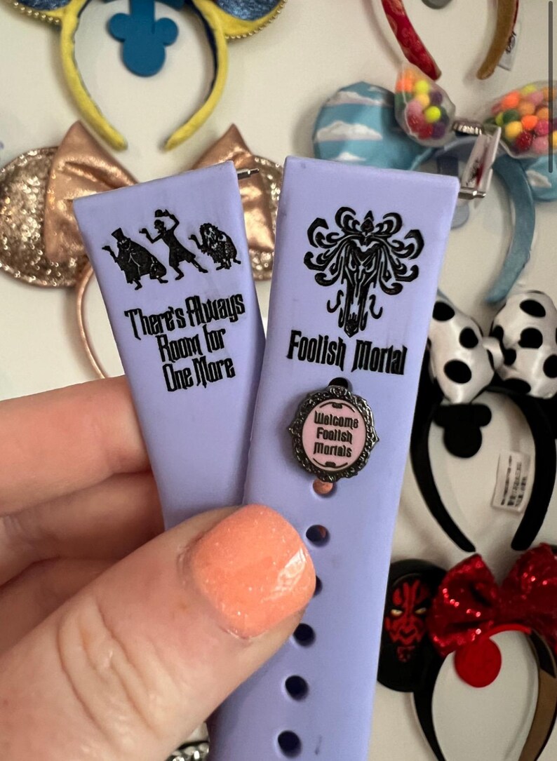 May include: Two purple silicone watch bands with black text and images. The band on the left says "There's Always Room for One More" and has a silhouette of three ghosts. The band on the right says "Foolish Mortal" and has a silhouette of a ghost with a swirling design. A small silver charm with pink text that says "Welcome Foolish Mortals" is attached to the right band.