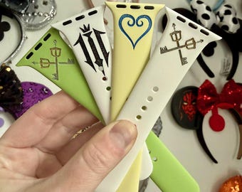 May include: A hand holds five colorful Apple Watch bands. The bands are lime green, white, and yellow, each with a unique black design. The background features various decorative headbands with bows and glitter.
