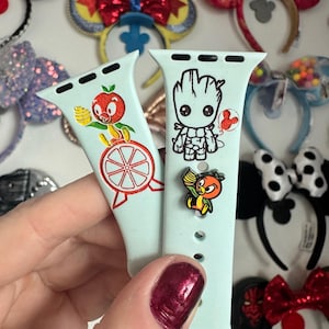 May include: Two light blue Apple Watch bands with different designs. One band features a cartoon orange with a red wheel and the other band features a cartoon Groot from Guardians of the Galaxy.