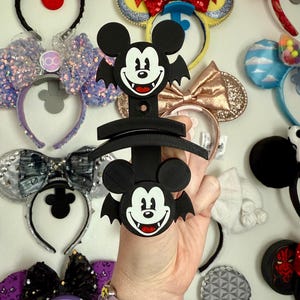 May include: A black Mickey Mouse-themed ear holder with a bat-like design. The holder features two Mickey Mouse faces with bat wings. The background shows various decorative headbands with bows and sequins.