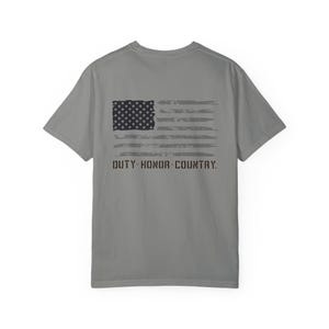 Puede incluir: Camiseta gris con un diseño desgastado de la bandera estadounidense en la espalda. La bandera es en tonos grises y negros, con el texto "DUTY HONOR COUNTRY" debajo. La camiseta es de cuello redondo.