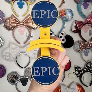 May include: A yellow display stand with two blue circular plaques that read "EPIC" in silver lettering. The stand is held in front of a wall covered in various decorative headbands with mouse ears.