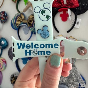 May include: A light blue Apple Watch band with a white background and the text "Welcome Home" in blue glitter. The band has a Mickey Mouse head design with a blue and green globe in the centre. There is a blue Mickey Mouse head charm with a gold "F" in the centre.