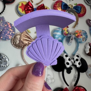 May include: Purple plastic headband holder shaped like a seashell. The holder has a curved top and a flat base. It is designed to hold headbands upright.