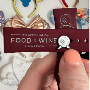 May include: A burgundy red watch band with a silver inscription that reads "INTERNATIONAL FOOD & WINE FESTIVAL". A white and green enamel pin with a sphere design is attached to the band.
