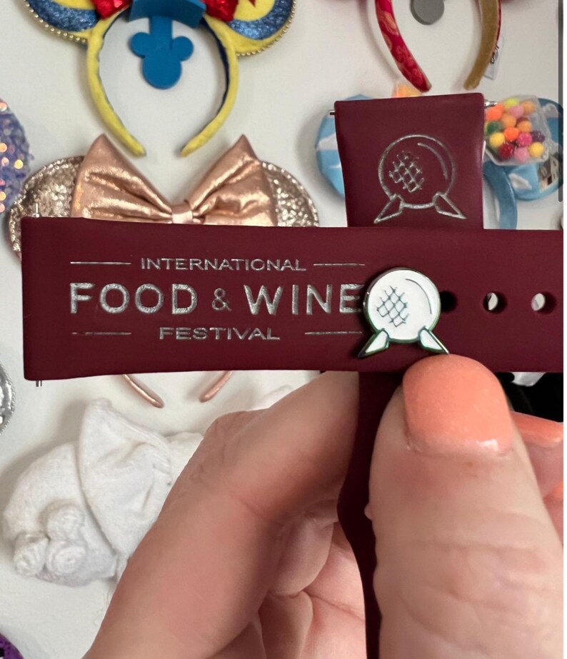 May include: A burgundy red watch band with a silver inscription that reads "INTERNATIONAL FOOD & WINE FESTIVAL". A white and green enamel pin with a sphere design is attached to the band.