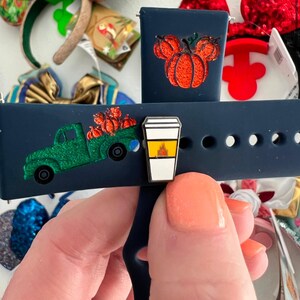 May include: A blue silicone watch band with two interchangeable charms. One charm is a green truck with pumpkins and the other is a pumpkin-shaped Mickey Mouse head. The charms are attached to the band with a metal pin.