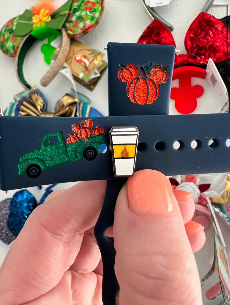 May include: A blue silicone watch band with two interchangeable charms. One charm is a green truck with pumpkins and the other is a pumpkin-shaped Mickey Mouse head. The charms are attached to the band with a metal pin.