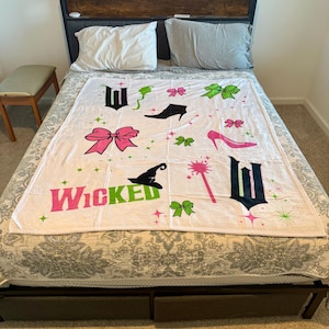 Peut inclure: Une couverture blanche avec le mot "Wicked" en rose et vert, ainsi que divers graphiques dont un chapeau de sorcière, des talons hauts, des nœuds et une baguette magique. La couverture est posée sur un lit.