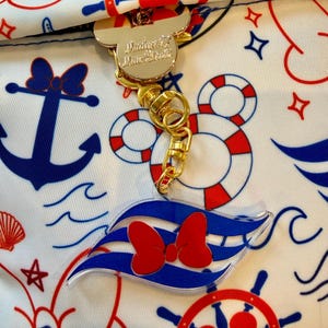 Könnte beinhalten: Ein goldfarbener Mickey-Mouse-Schlüsselanhänger mit einem transparenten Acryl-Anhänger mit roter Schleife und blauem Wellenmuster. Der Schlüsselanhänger ist an einem weißen Stoff mit nautischen Mustern in Rot und Blau befestigt.