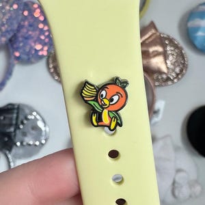 May include: A yellow Apple Watch band with a small orange bird charm attached. The charm is of a cartoon bird holding a paintbrush.
