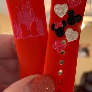 May include: Two red silicone watch bands with different designs. One band features a pink and white castle with a bow, the other band has hearts and Mickey Mouse heads with phrases like "XOXO" and "Be Mine".