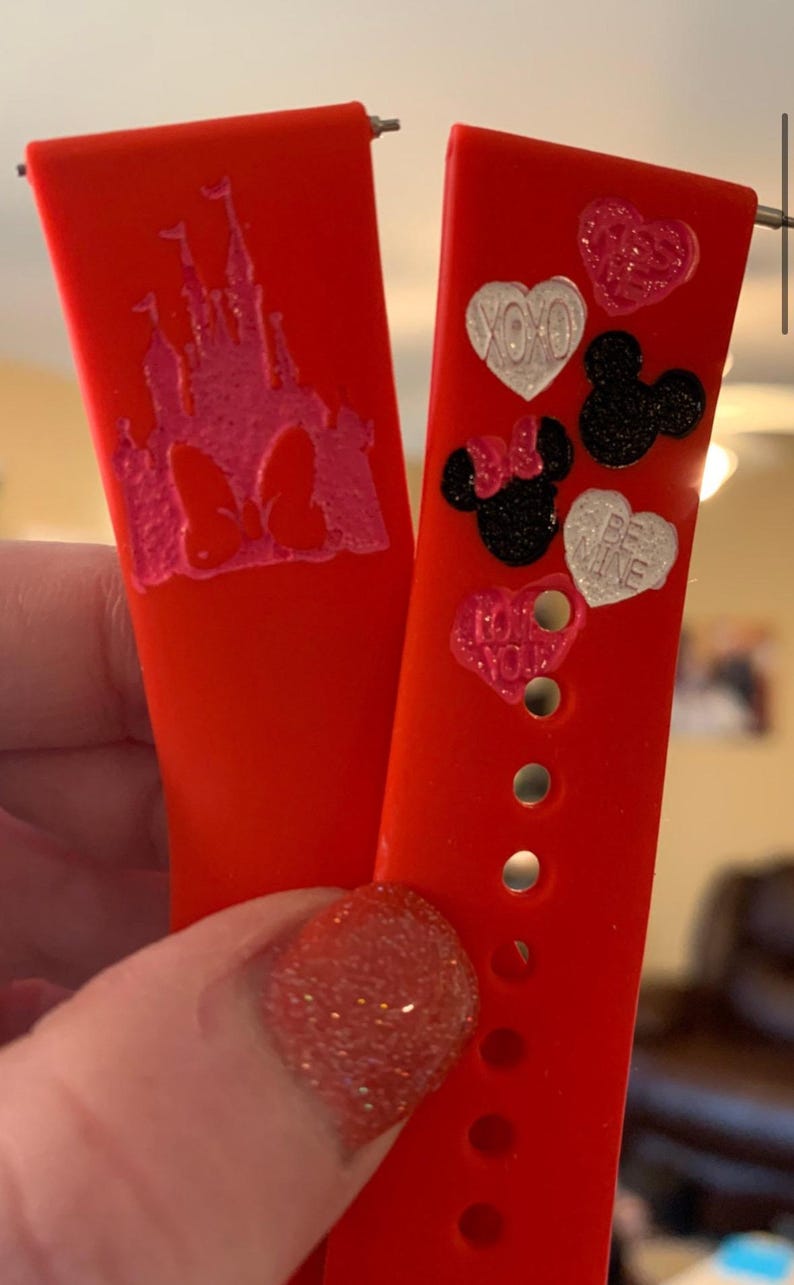 May include: Two red silicone watch bands with different designs. One band features a pink and white castle with a bow, the other band has hearts and Mickey Mouse heads with phrases like "XOXO" and "Be Mine".