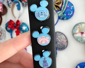 Puede incluir: Tres dijes en forma de Mickey Mouse de color azul claro con diferentes diseños de edificios en una correa de reloj negra. Los dijes tienen un borde blanco. Otros elementos decorativos están en el fondo.