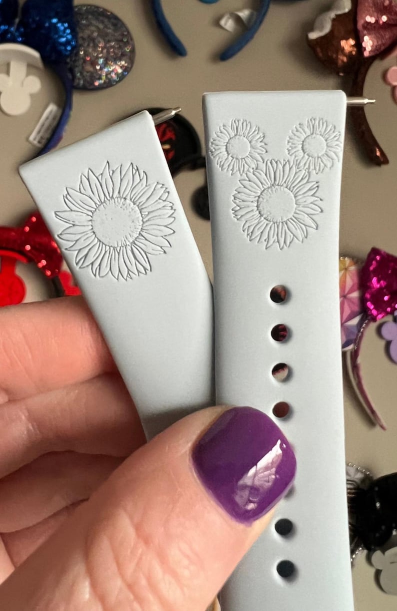 May include: Two light blue silicone watch bands with a white sunflower design. The band on the right has three sunflowers arranged in a Mickey Mouse shape.