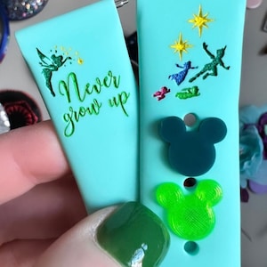 May include: Two turquoise blue watch bands with different designs. One band features the text "Never grow up" with a green fairy silhouette. The other band has a green silhouette of Peter Pan flying with Tinkerbell and the words "Second Star to the Right".