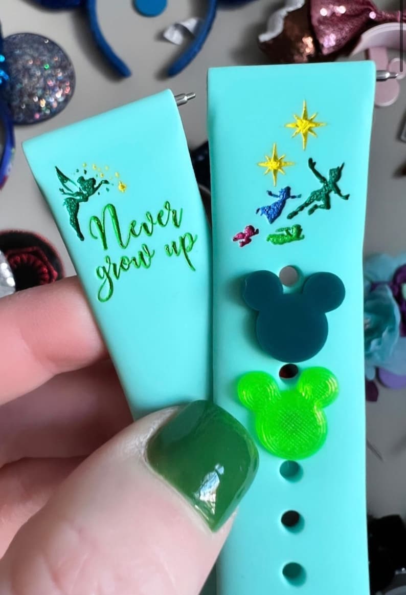 May include: Two turquoise blue watch bands with different designs. One band features the text "Never grow up" with a green fairy silhouette. The other band has a green silhouette of Peter Pan flying with Tinkerbell and the words "Second Star to the Right".