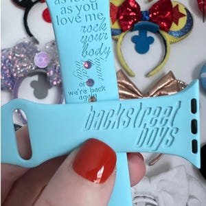 以下が含まれることがあります： 「as long as you love me, rock your body」と「Backstreet Boys」の文字が刻まれた、水色の時計バンド。様々な装飾的なヘアバンドを背景にしています。