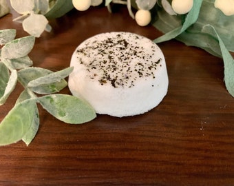 May include: A round, white bath bomb with a dark green speckled topping. The bath bomb sits on a dark brown wooden surface, with green and white decorative leaves and berries in the background.
