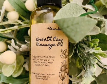 May include: A small, clear glass bottle with a black cap, labeled "Fleur de Breath Easy Massage Oil". The bottle contains a yellow liquid and is surrounded by green foliage.