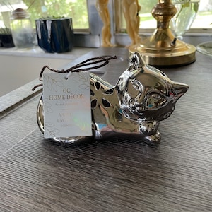 May include: Silver ceramic cat figurine with a cutout design. The figurine is a home decor item and is scented with vanilla and lavender.