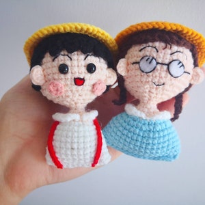 May include: Two crocheted dolls, each with a yellow hat and smiling face. One doll has black hair and a white shirt with red accents. The other doll has brown hair, glasses, and a blue top. Both dolls have rosy cheeks.