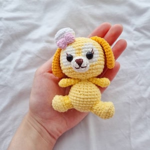 May include: A handmade yellow crochet puppy with a white and pink bow. The puppy has black eyes, a brown nose, and yellow ears. The crochet animal is held in a hand against a white background.
