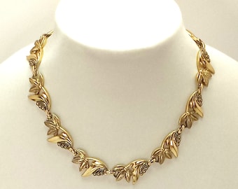 May include: A gold-toned necklace with a leaf-like design, displayed on a white mannequin. The necklace features repeating, stylized leaf shapes connected by small links. The leaves have a textured surface and are arranged in a symmetrical pattern.