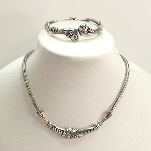 Op de afbeelding: Een zilveren ketting- en armbandset met een gedraaid touwontwerp en sierlijke details. De ketting heeft een gebogen vorm en de armband heeft een swirl-ontwerp.
