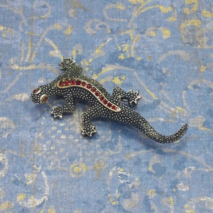 May include: A black lizard brooch with red rhinestone accents. The lizard is detailed with a textured finish and has a star-shaped design on its back.