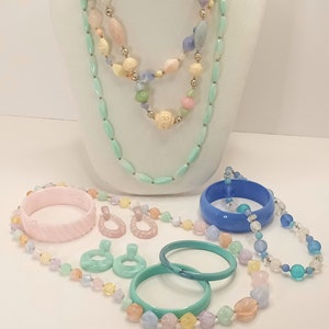 Peut inclure: Un ensemble de bijoux de couleur pastel comprenant un long collier à plusieurs rangs de perles, deux bracelets, une paire de boucles d'oreilles et un collier plus court. Les perles sont de formes et de tailles variées, notamment ovales, rondes et carrées. Les couleurs des perles sont le bleu clair, le rose, le vert et le jaune.
