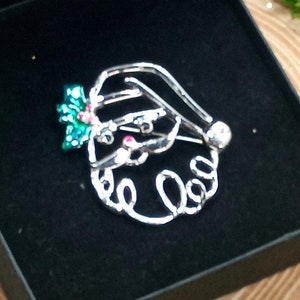 May include: A silver wire Santa Claus brooch with green and red holly accents. The brooch is displayed in a black velvet box.