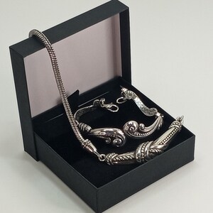 Op de afbeelding: Een zilveren ketting- en armbandset in een zwarte geschenkdoos. De ketting heeft een gevlochten ketting en de armband heeft een decoratief ontwerp.