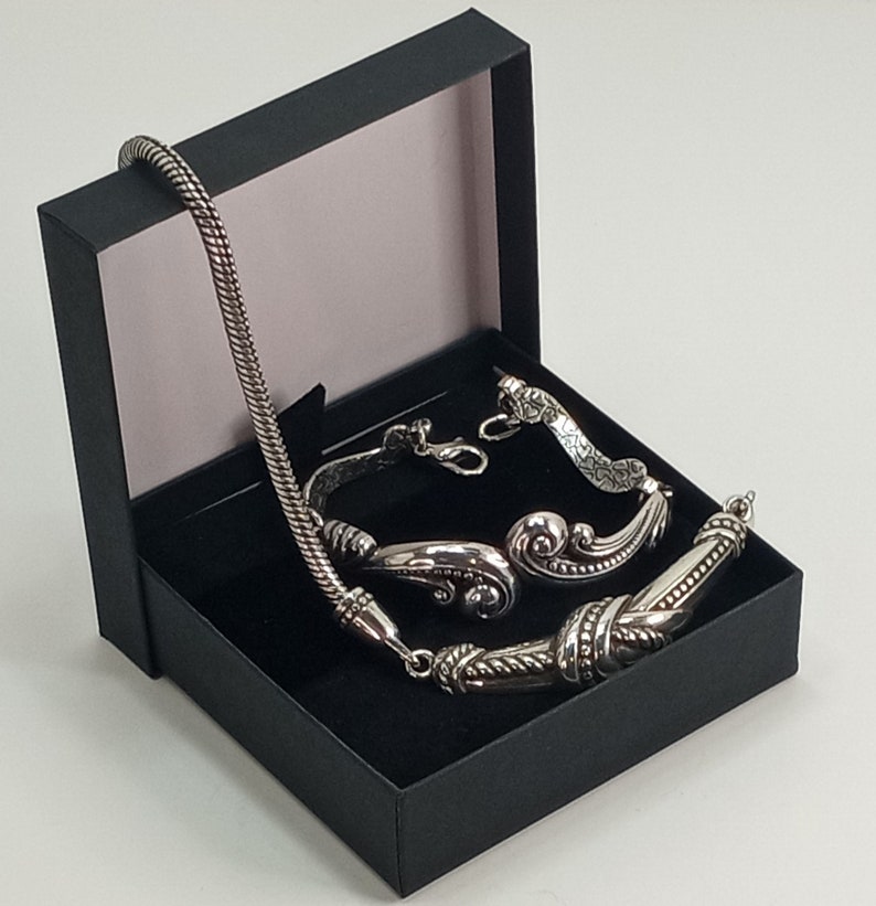 Op de afbeelding: Een zilveren ketting- en armbandset in een zwarte geschenkdoos. De ketting heeft een gevlochten ketting en de armband heeft een decoratief ontwerp.